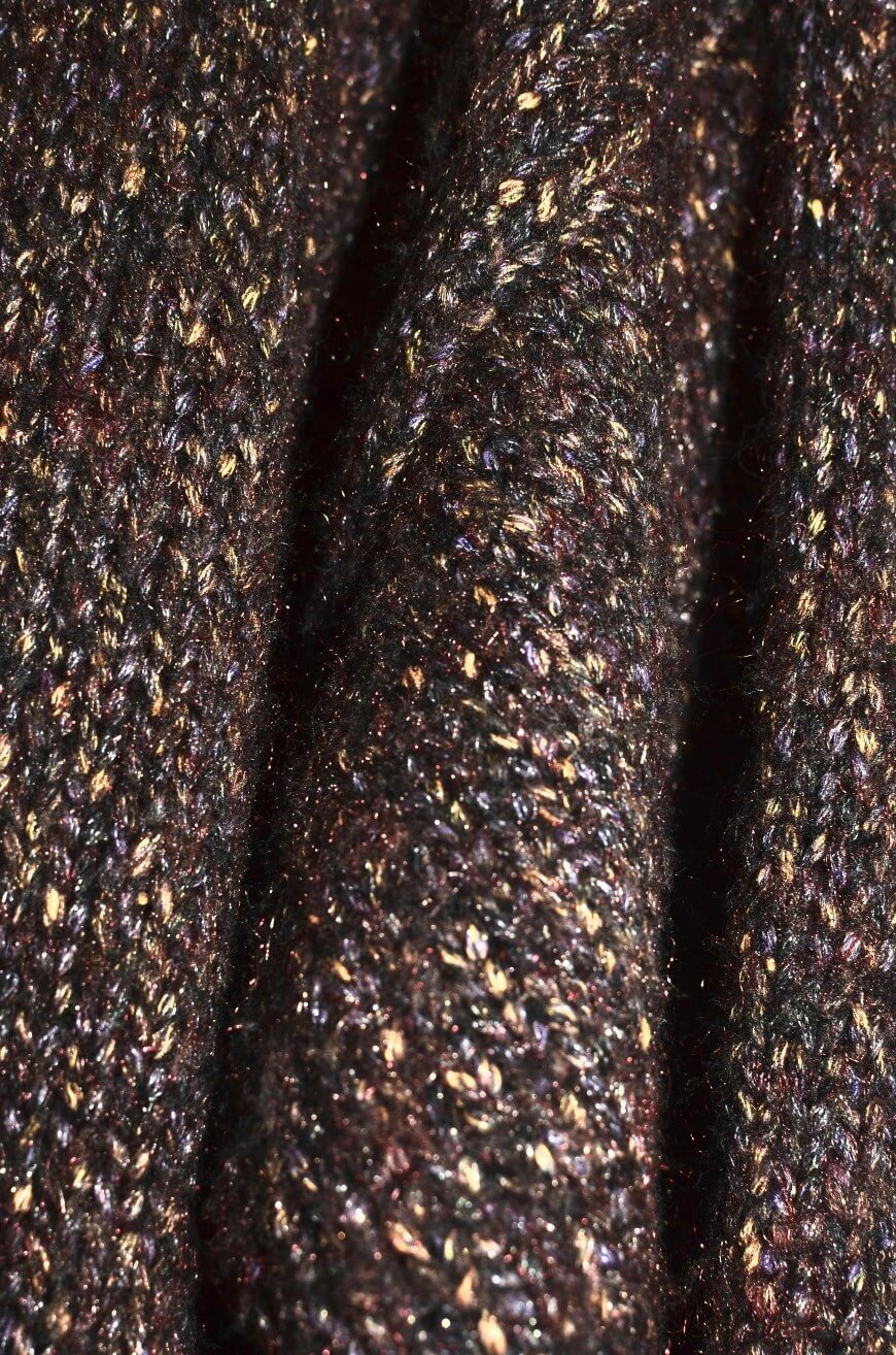 Close-up of a textured, knitted fabric with dark brown, black, and gold speckled threads, showing tightly woven stitches and a slightly shimmery appearance.