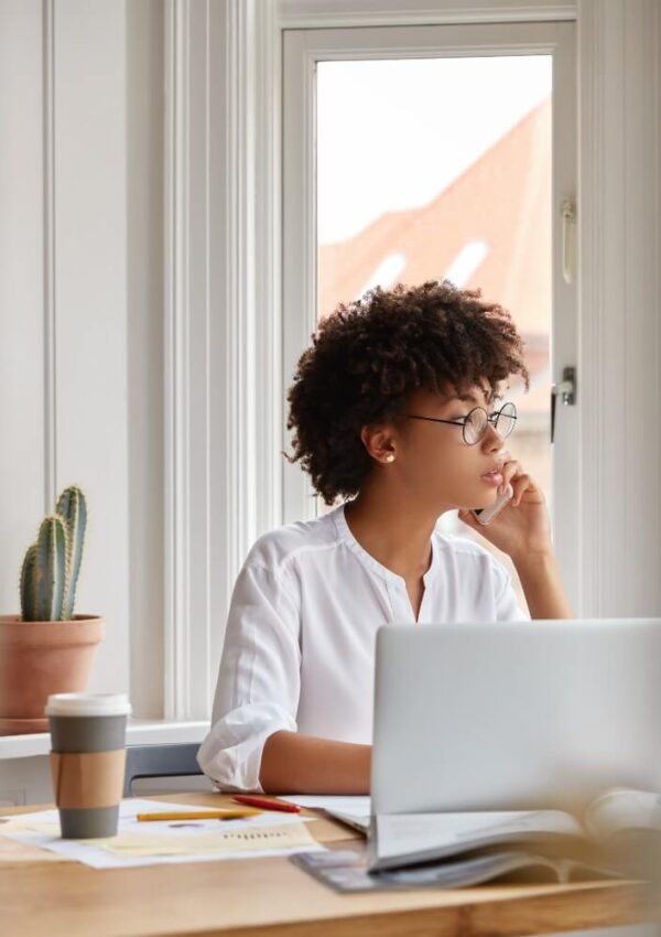 A woman with curly hair and glasses sits at a desk with a laptop, papers, and a coffee cup, looking thoughtfully out a window. A potted cactus is on the windowsill behind her.