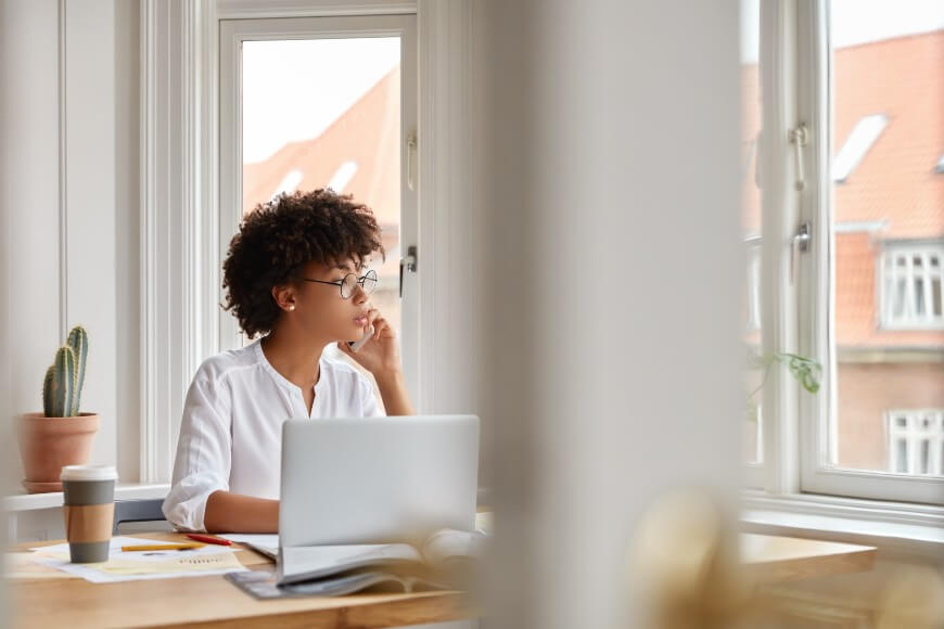 A woman with curly hair and glasses sits at a desk with a laptop, papers, and a coffee cup, gazing thoughtfully out a large window. A potted cactus is on the windowsill beside her.