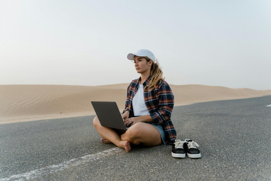 A woman sits barefoot on a deserted road in the desert, using a laptop. She wears a plaid shirt, denim shorts, and a white cap. Her shoes are placed beside her on the road, and sand dunes are visible in the background.