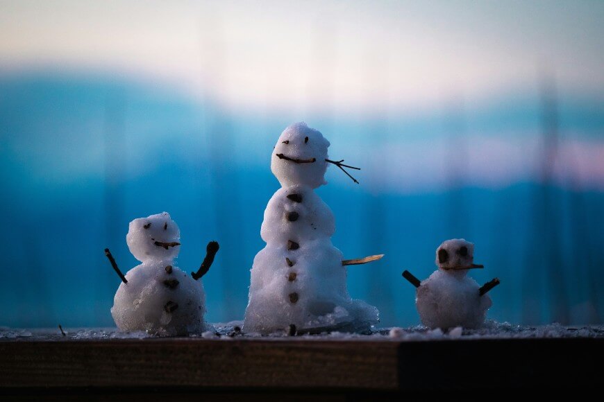 Three small snowmen with stick arms and smiling faces stand together on a wooden surface, with a soft, out-of-focus blue and pink background suggesting dusk or dawn.