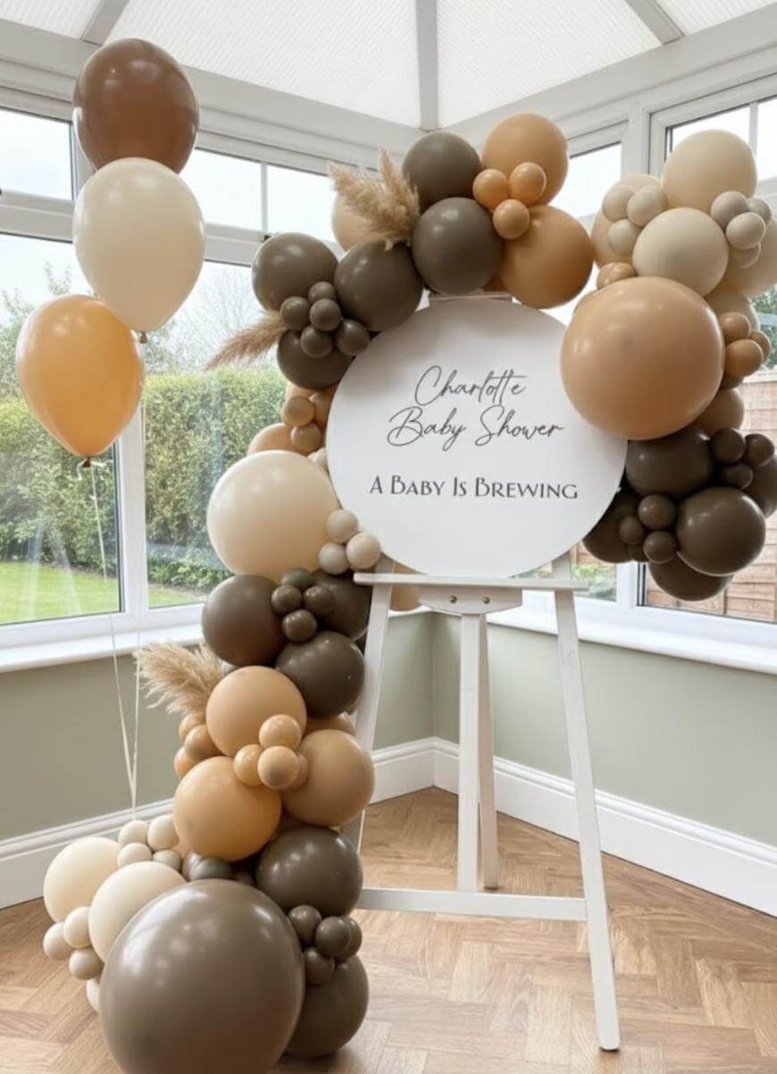 A baby shower display with brown, beige, and cream balloons arranged around a sign on an easel that reads Charlotte Baby Shower - A Baby Is Brewing. The setup is in a bright room with large windows.