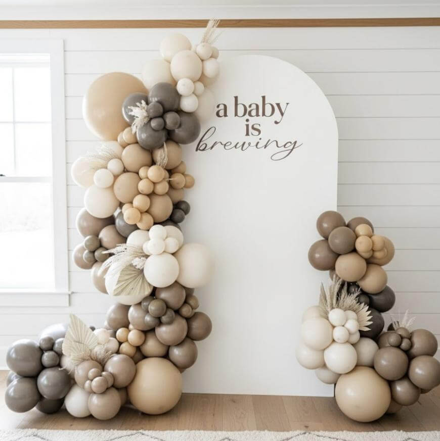 A white decorative backdrop with the words “a baby is brewing,” surrounded by clusters of beige, brown, and cream balloons and pampas grass, set in a bright room with white paneled walls.