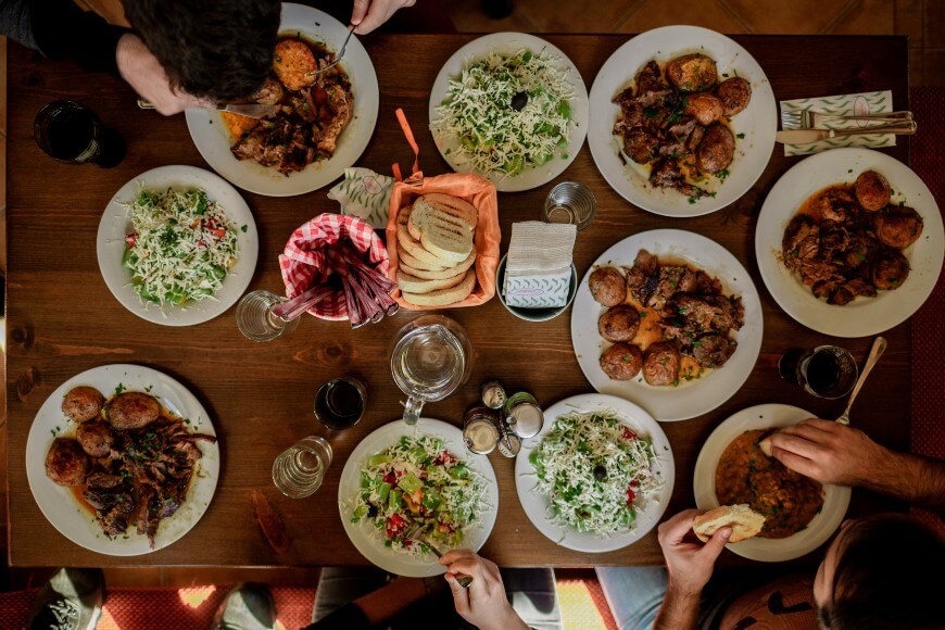 A group of people share a meal at a wooden table set with plates of salad, roasted potatoes, meat, a basket of bread, glasses of dark beverage, and a water pitcher. Hands are reaching for food and eating.