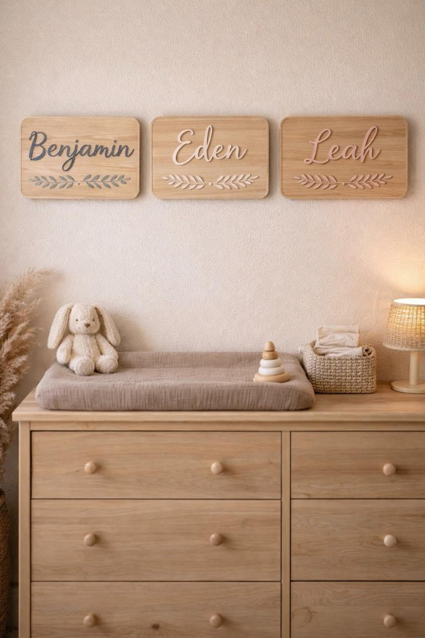 A wooden dresser with a changing pad, plush bunny, and lamp on top. Above it, three wooden name plaques read Benjamin, Eden, and Leah. The decor includes soft, neutral tones and dried pampas grass.