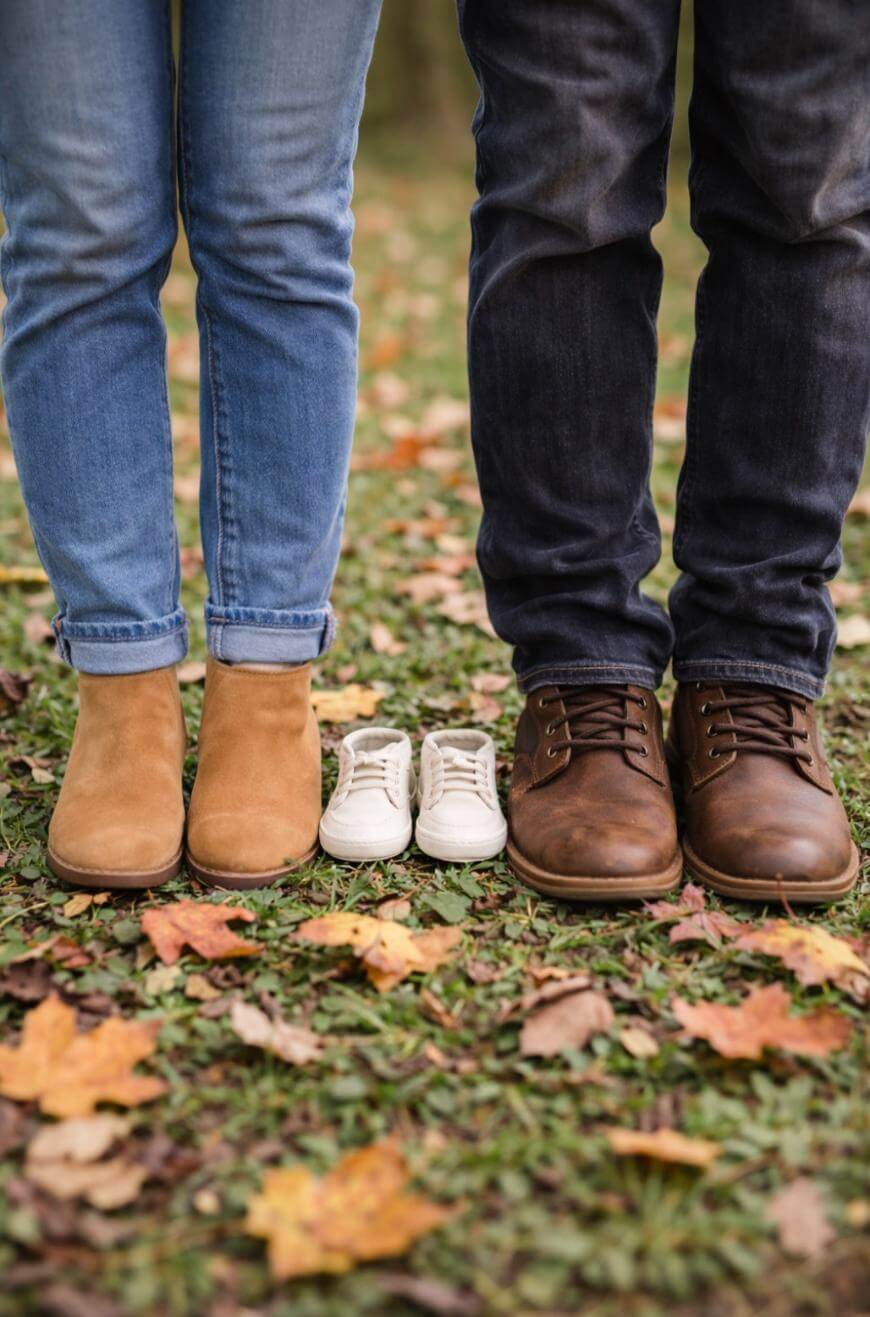 A woman and man stand side by side on grass with fallen autumn leaves, wearing jeans and boots. Between them is a small pair of baby shoes, suggesting an upcoming addition to their family.
