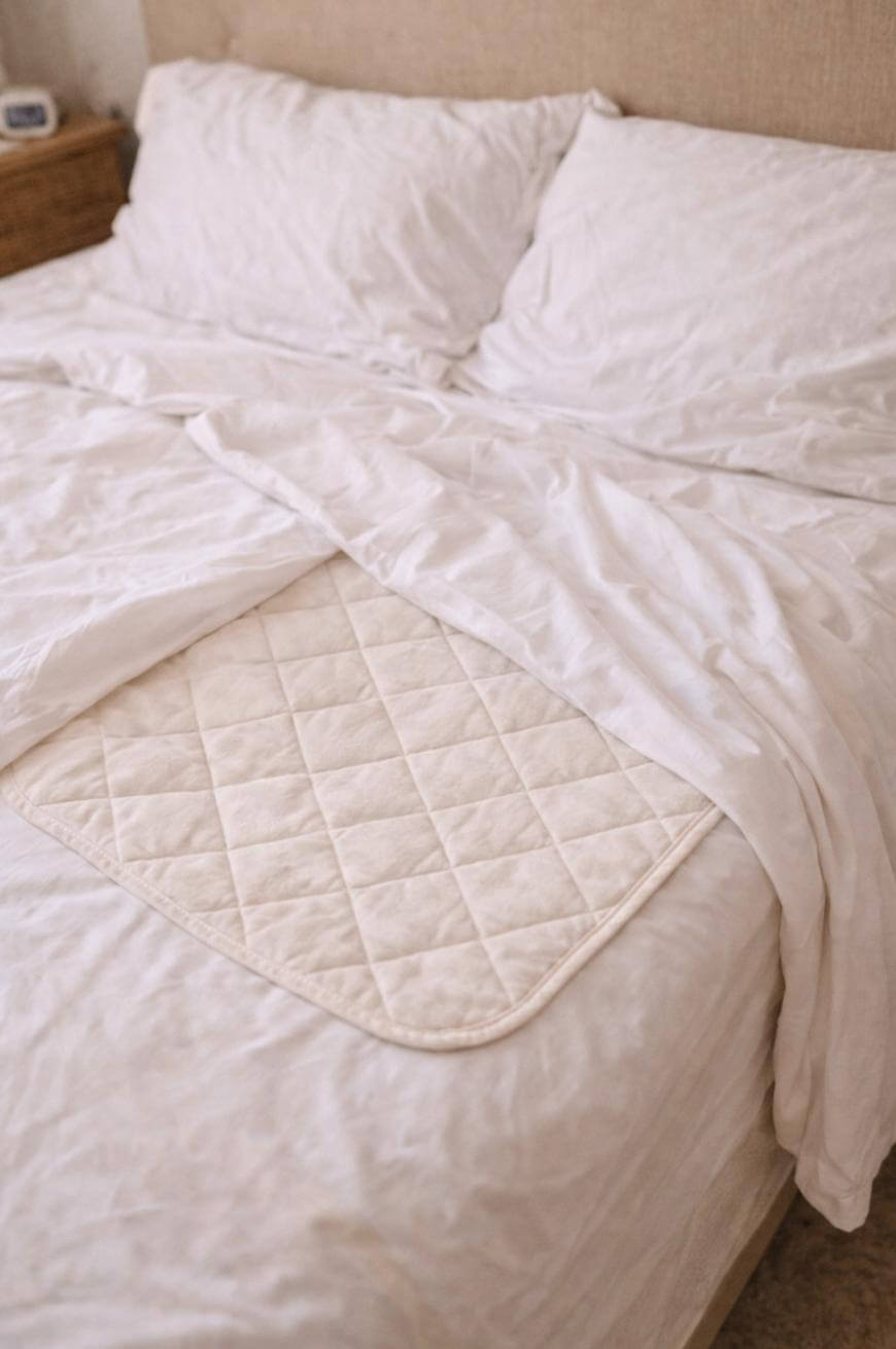 A neatly made bed with white sheets and pillows, featuring a quilted, beige waterproof pad placed on top of the sheet near the center of the bed.