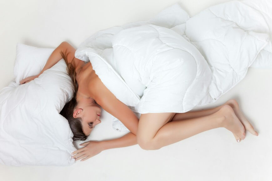 A woman lies on her side in a fetal position, covered with a white blanket on a white bed, resting her head on a pillow.