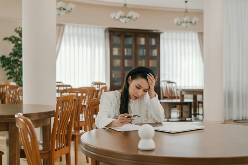 A young woman sits alone at a round wooden table in a bright room, looking stressed as she holds her head with one hand while studying papers. A closed laptop and pen are on the table beside her.