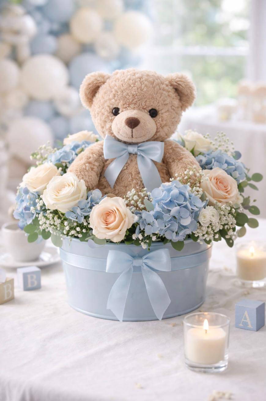 A plush teddy bear with a blue bow sits in a light blue hatbox filled with cream and blue flowers. The box is tied with a matching ribbon. Lit candles and letter blocks decorate the soft, light-filled background.