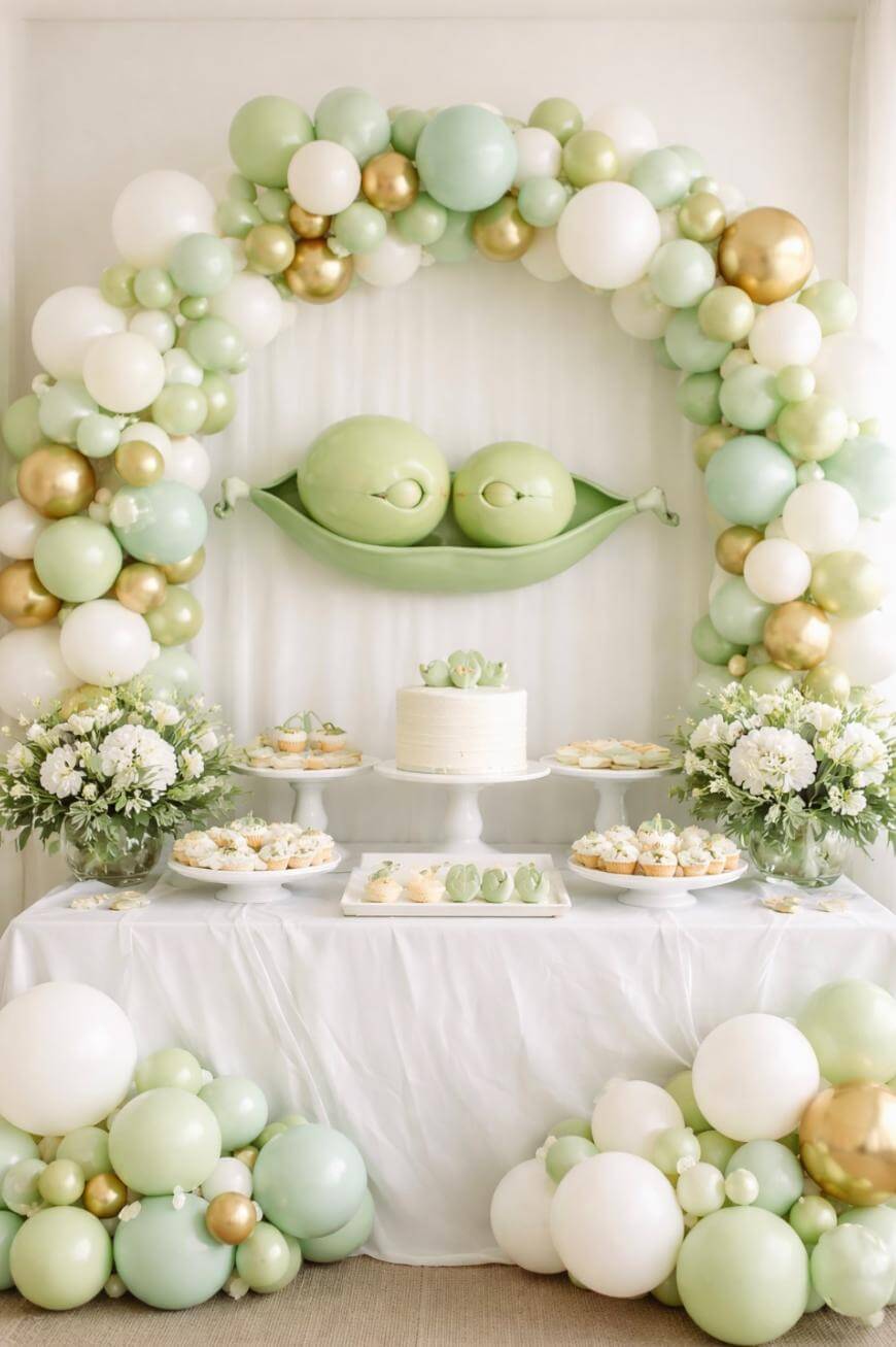 A dessert table decorated with white, green, and gold balloons, a cake, sweet treats, and flower arrangements, with a “two peas in a pod” theme featuring a pea pod decoration above the table.