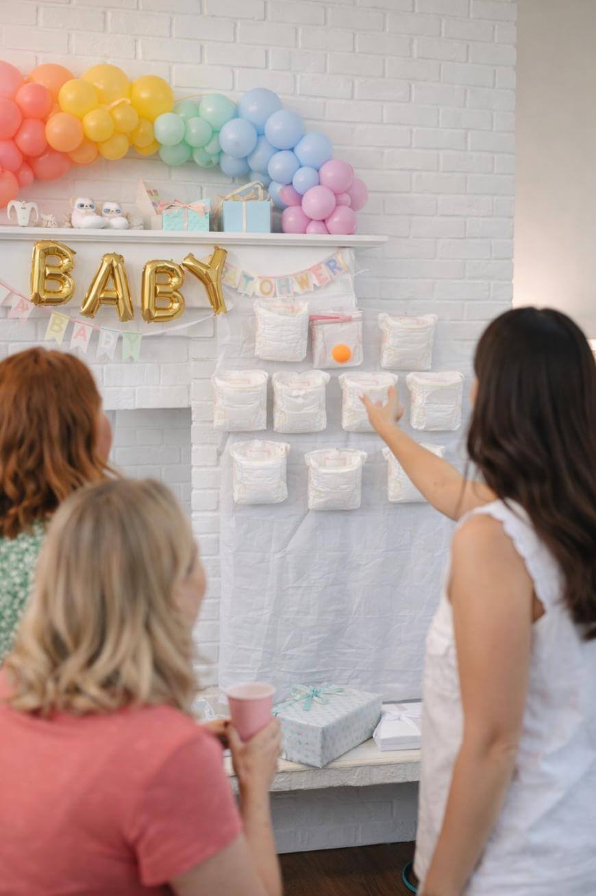 Three women play a baby shower game involving diapers attached to a wall. One woman is tossing a ping pong ball at the diapers. Colorful balloons and gold BABY balloons decorate the white brick wall.
