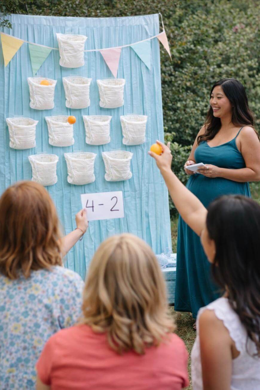 A woman in a teal dress smiles as guests throw balls at hanging diaper targets, with a sign reading 4-2 beneath them. The scene is outdoors with colorful bunting and people seated facing the game.