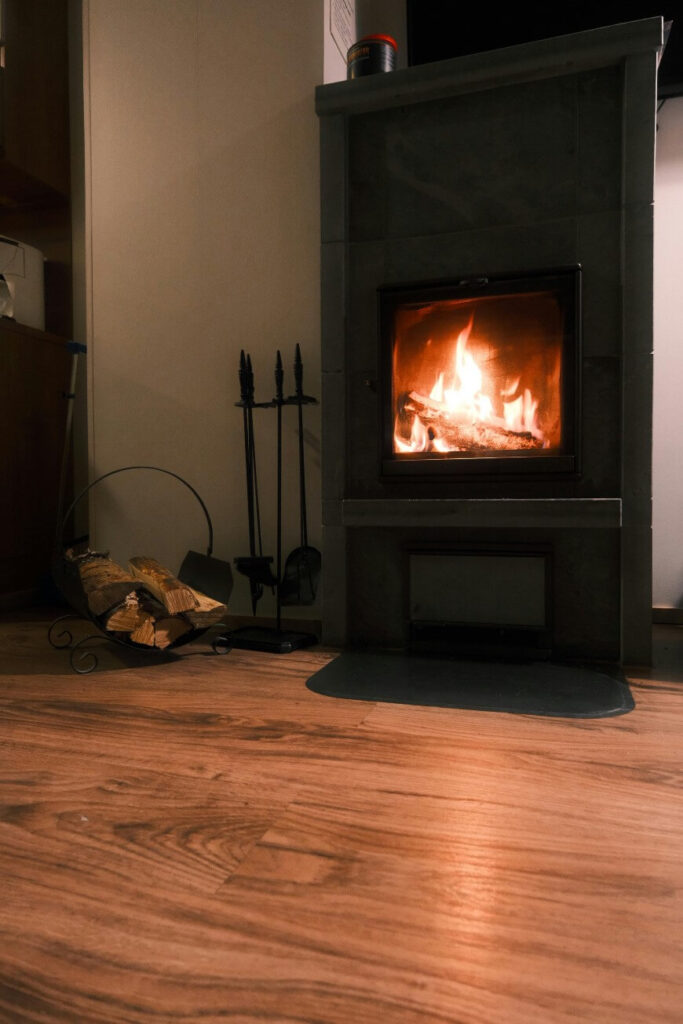 A cozy wood-burning stove with a fire inside, set against a wall on wooden flooring. A small rack holding firewood and fireplace tools sits nearby. The warm glow of the fire lights the room.