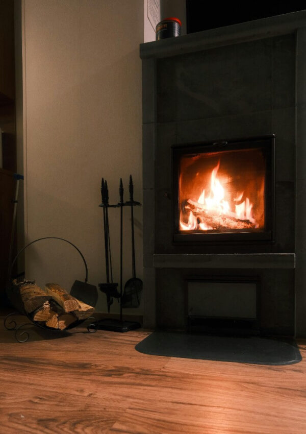 A cozy indoor scene with a wood-burning stove emitting a warm fire, a set of fireplace tools, and a basket of chopped firewood on a wooden floor beside the stove.