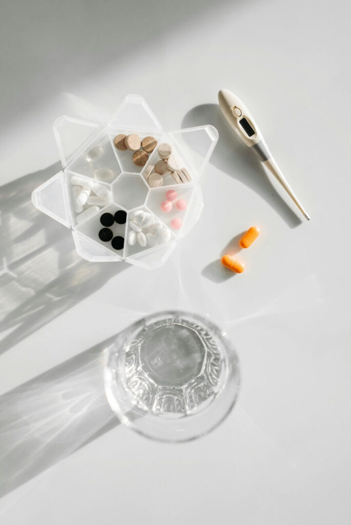 A pill organizer with assorted pills, a digital thermometer, two orange capsules, and a glass of water on a white surface.