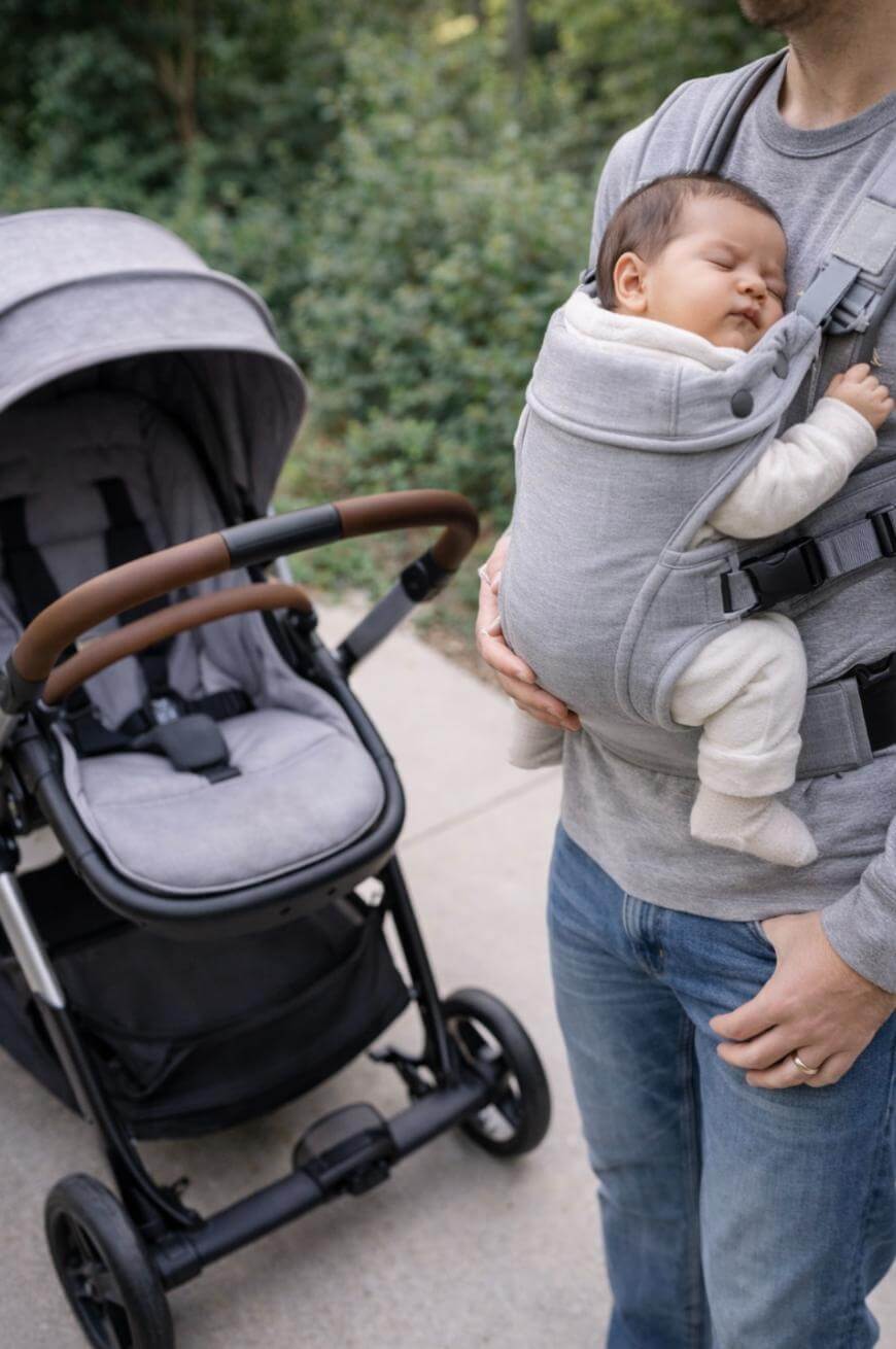 A man stands outdoors holding a sleeping baby in a gray carrier on his chest. Next to them is an empty gray stroller. Trees and greenery are visible in the background.