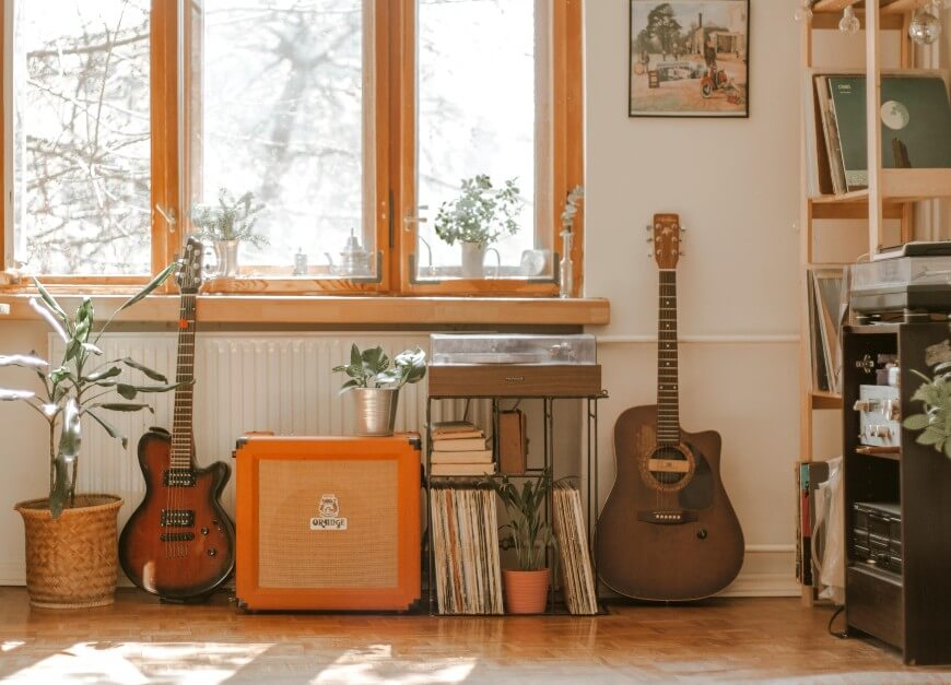 Two guitars and an orange amplifier sit beneath a window with potted plants, surrounded by vinyl records, turntable, and shelves of music equipment in a sunlit, cozy room.