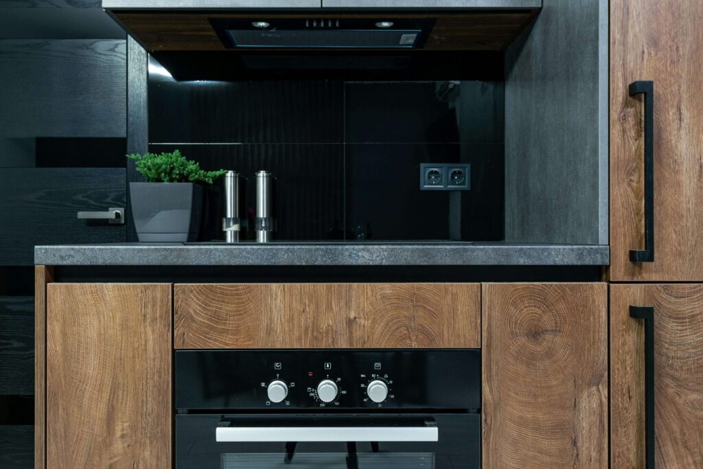Modern kitchen with wooden cabinets, a built-in oven, black glass backsplash, potted green plant, and two metallic salt and pepper shakers on the countertop. Electric sockets are visible on the back wall.