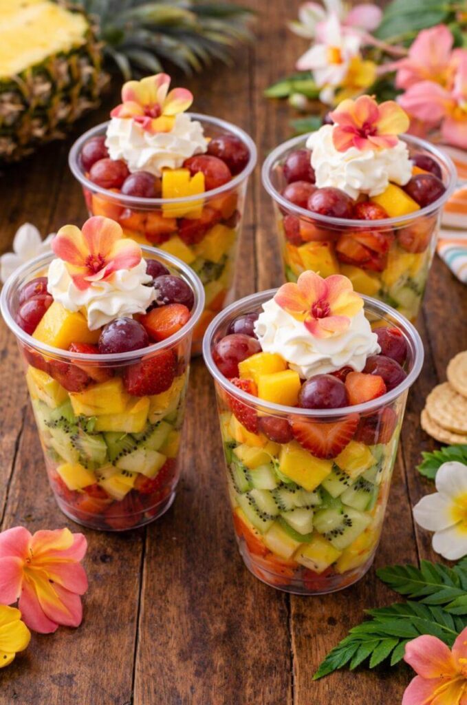 Four clear plastic cups filled with colorful layers of chopped fruit, topped with whipped cream and decorative edible flowers, are displayed on a wooden table with tropical flowers and leaves around them.