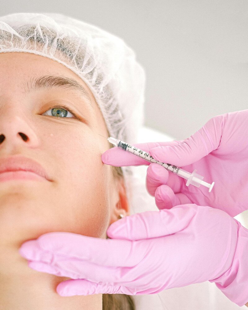 A person wearing a hair cover receives a cosmetic injection near the eye from someone wearing pink gloves, who is holding a syringe and gently supporting the persons face.