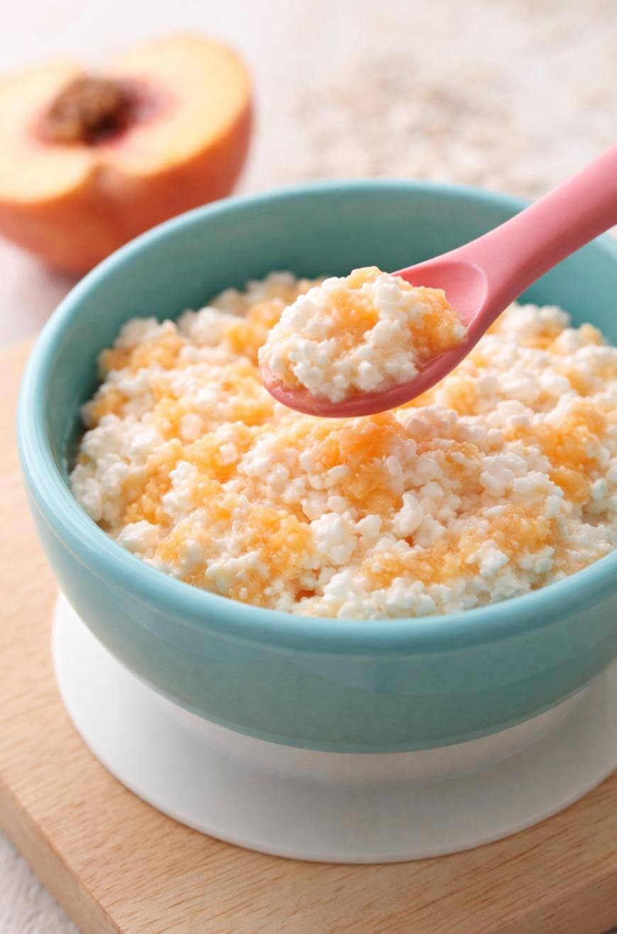 A blue bowl filled with cottage cheese topped with peach puree, with a pink spoon lifting a spoonful. A halved peach is blurred in the background, all set on a wooden surface.