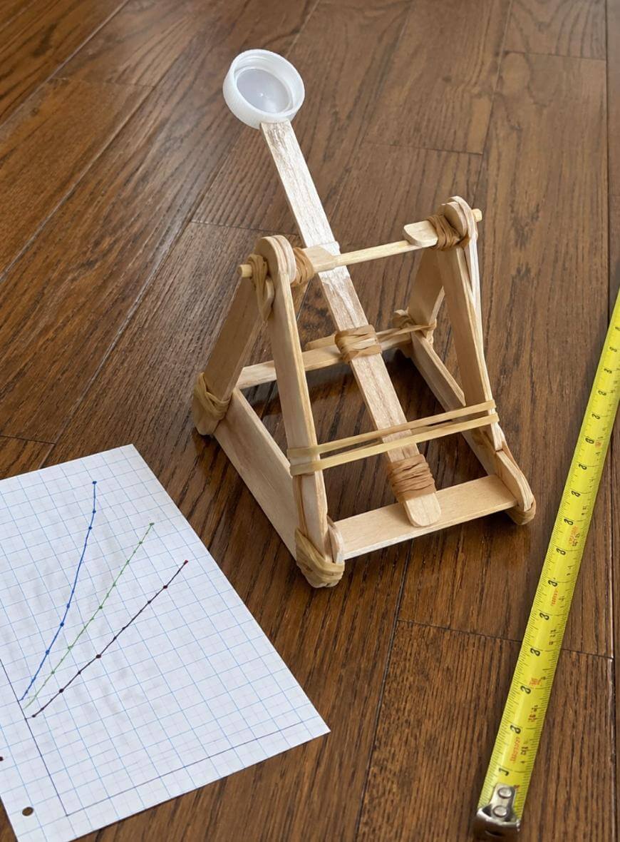 A small homemade catapult made from popsicle sticks, rubber bands, and a plastic spoon sits on a wooden floor next to a tape measure and a sheet of graph paper with hand-drawn lines.