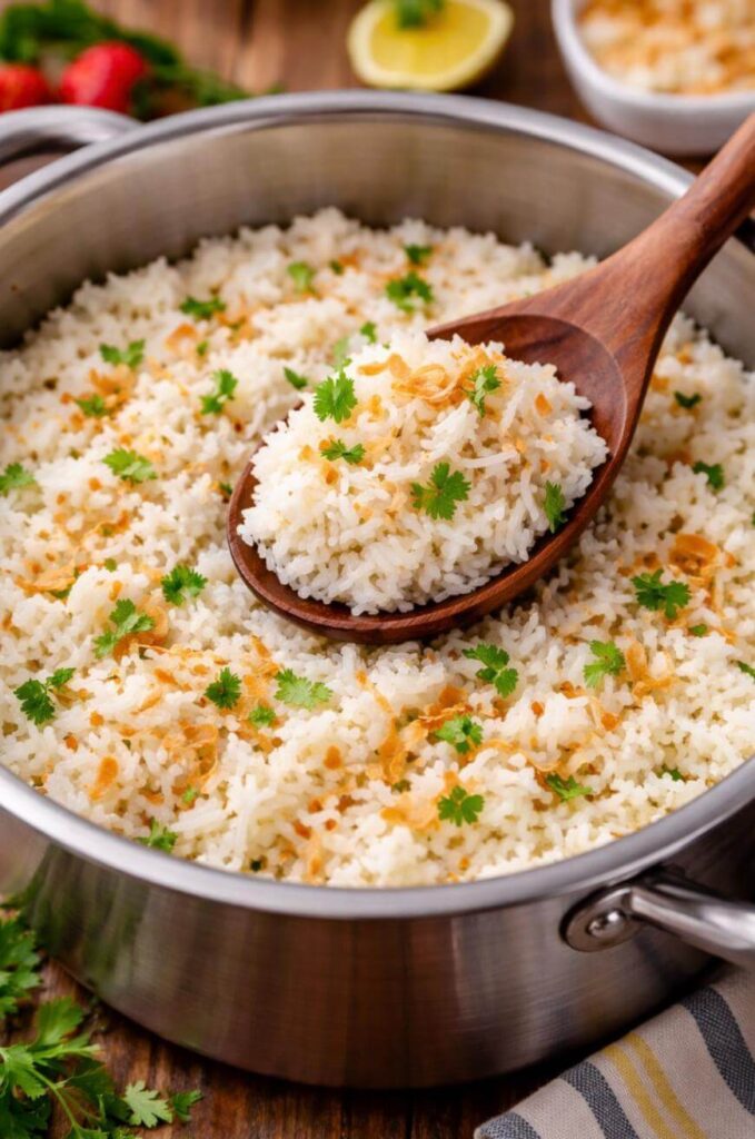 A pot of cooked white rice garnished with chopped herbs and crispy fried onions, with a wooden spoon lifting a serving from the pot.