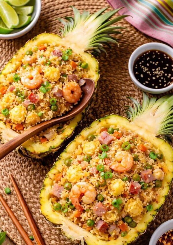 Two pineapple halves filled with fried rice, shrimp, peas, carrots, and diced ham, garnished with green onions. A wooden spoon, lime wedges, dipping sauce, and chopsticks are nearby on a woven placemat.