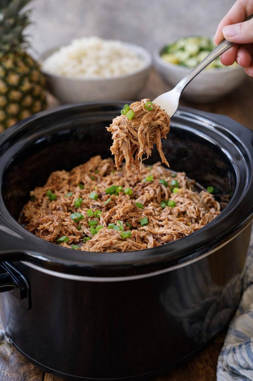 Shredded pork topped with chopped green onions in a black slow cooker, with a hand holding a forkful above. Bowls of rice and pineapple are in the background.