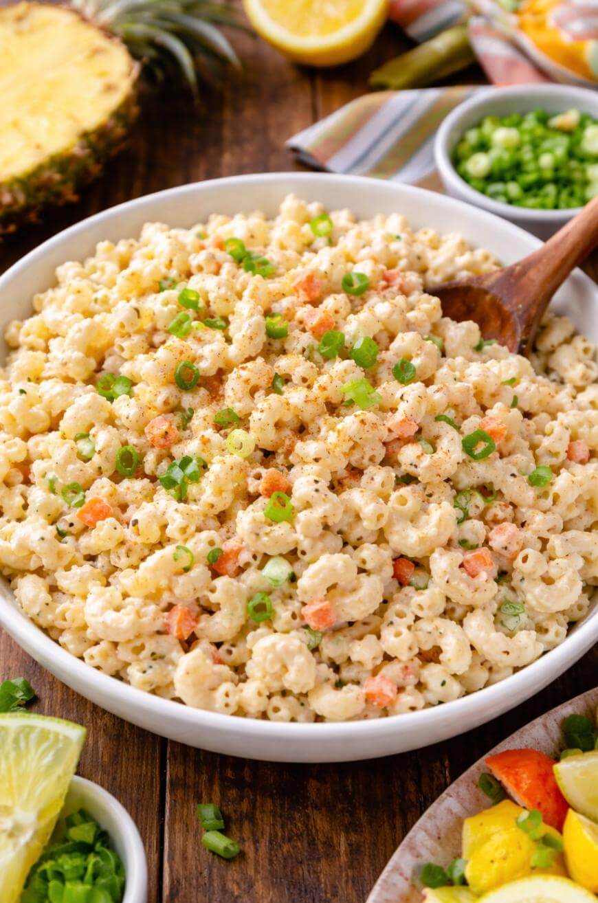 A bowl of creamy macaroni salad with chopped green onions and small carrot pieces, served with a wooden spoon. Fresh lemon and lime wedges, chopped green onions, and pineapple are visible in the background.