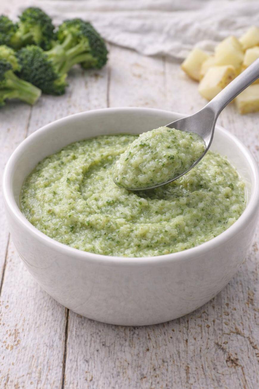 A white bowl filled with creamy green broccoli puree sits on a wooden surface, with a spoonful lifted above the bowl. Pieces of broccoli and diced potatoes are in the background.