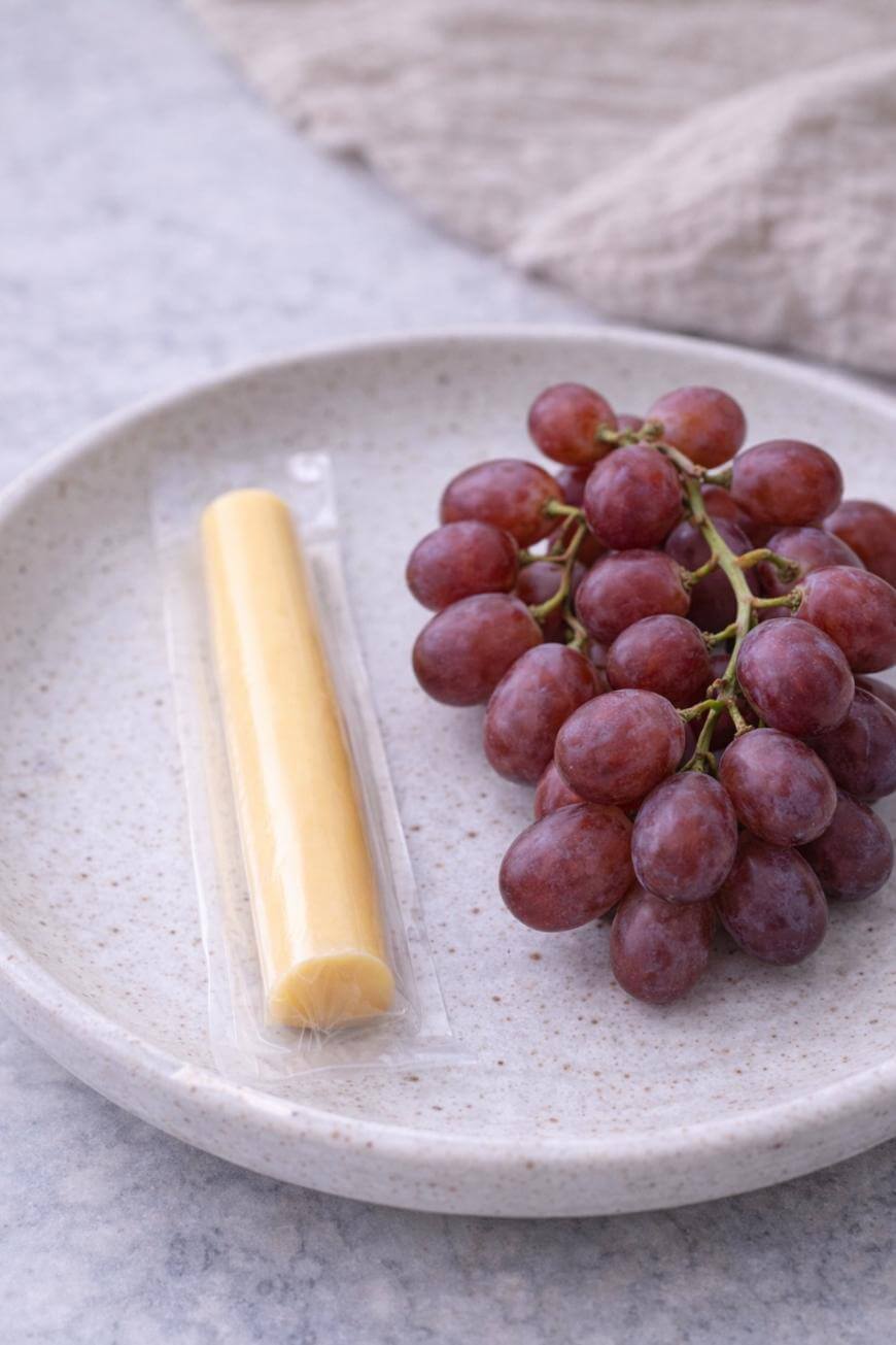 A bunch of red grapes and a packaged stick of cheese on a speckled white plate, placed on a light-colored surface.