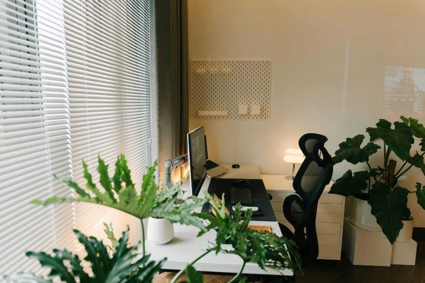 Minimalist home office with a black mesh chair, white desk, computer monitor, keyboard, mouse, small lamp, potted plants, and large window with blinds letting in natural light.