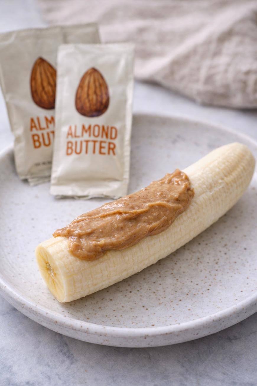A peeled banana on a speckled plate with almond butter spread on top. In the background, two almond butter packets are visible.