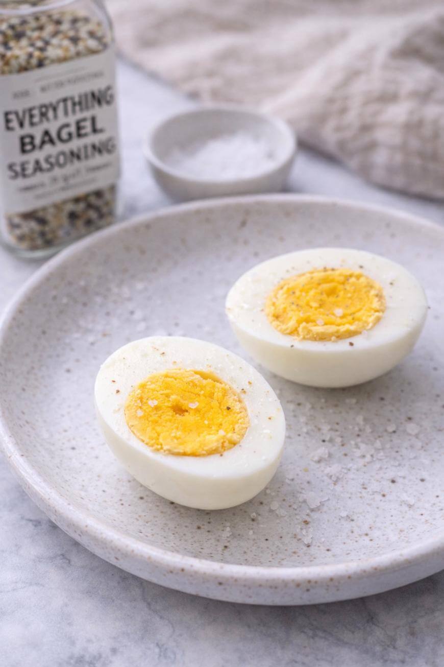 A hard-boiled egg cut in half sits on a speckled plate, sprinkled lightly with Everything Bagel seasoning. A jar of seasoning and a small bowl of salt are in the background.