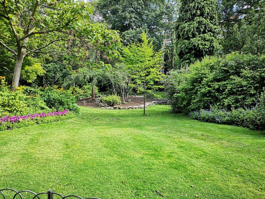 A lush green garden with a well-manicured lawn, surrounded by various trees, shrubs, and colorful flowerbeds. A small stone-bordered area with bare soil sits near the center, beneath leafy trees.
