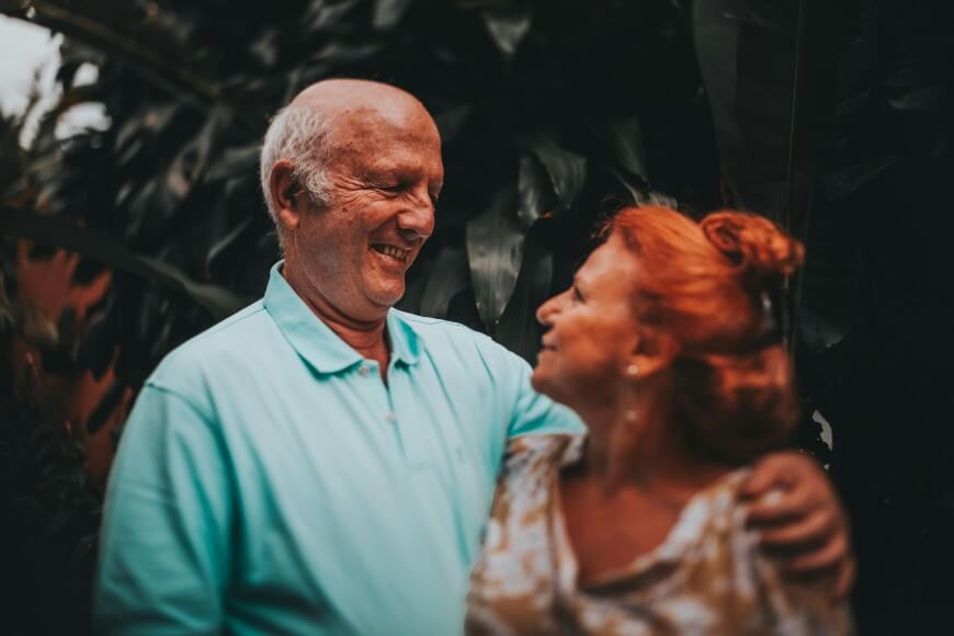 An older man and woman stand outdoors, smiling warmly at each other. The man wears a light blue polo shirt and has his arm around the woman, who has red hair and wears a patterned blouse. Green foliage is in the background.