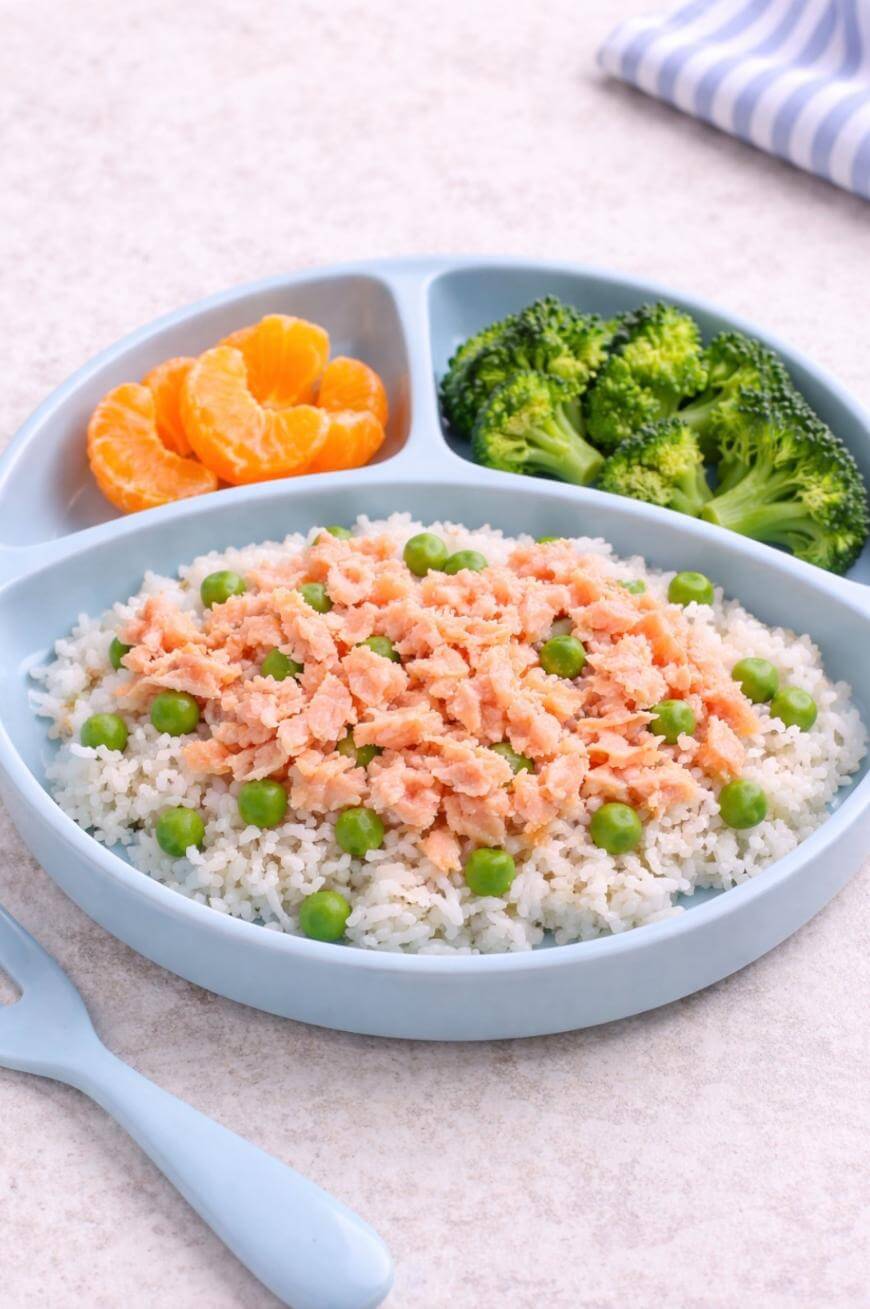 A blue divided plate with white rice topped with peas and flaked salmon, broccoli florets, and orange slices. A light blue fork is beside the plate, and a striped napkin is in the background.