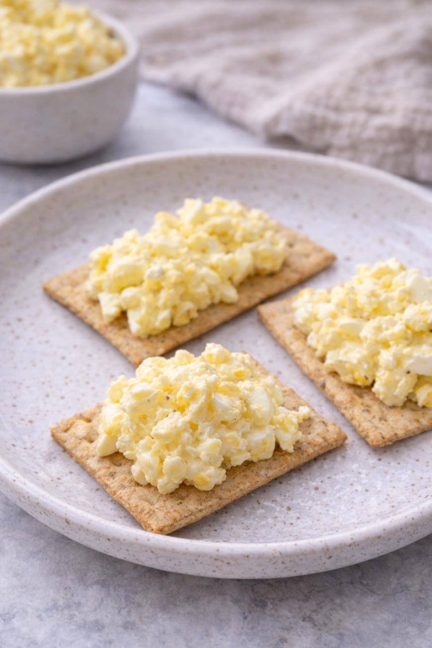 Three crackers topped with a creamy egg salad are arranged on a speckled ceramic plate, with a bowl of additional egg salad and a beige cloth in the background.