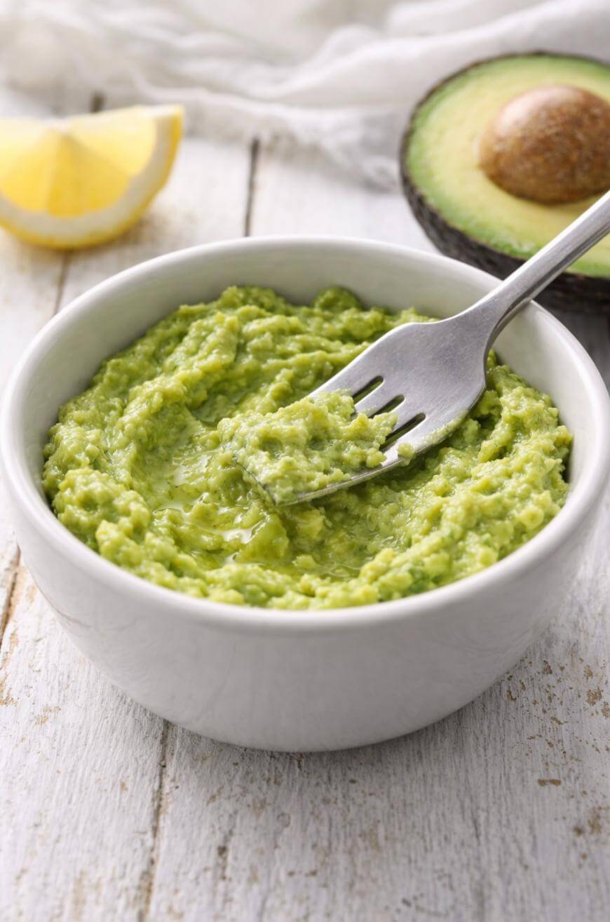 A white bowl filled with mashed avocado sits on a white wooden surface. A fork rests in the bowl. A lemon wedge and half an avocado are visible in the background.