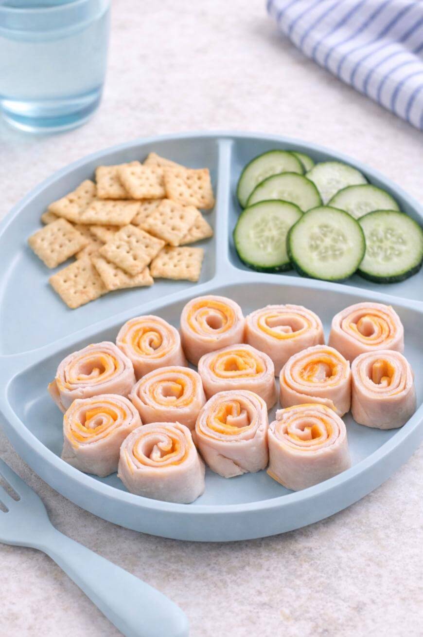 A divided plate with rolled turkey and cheese pinwheels, round cucumber slices, and square crackers. A glass of water and a striped cloth napkin are in the background. A blue fork is on the table.