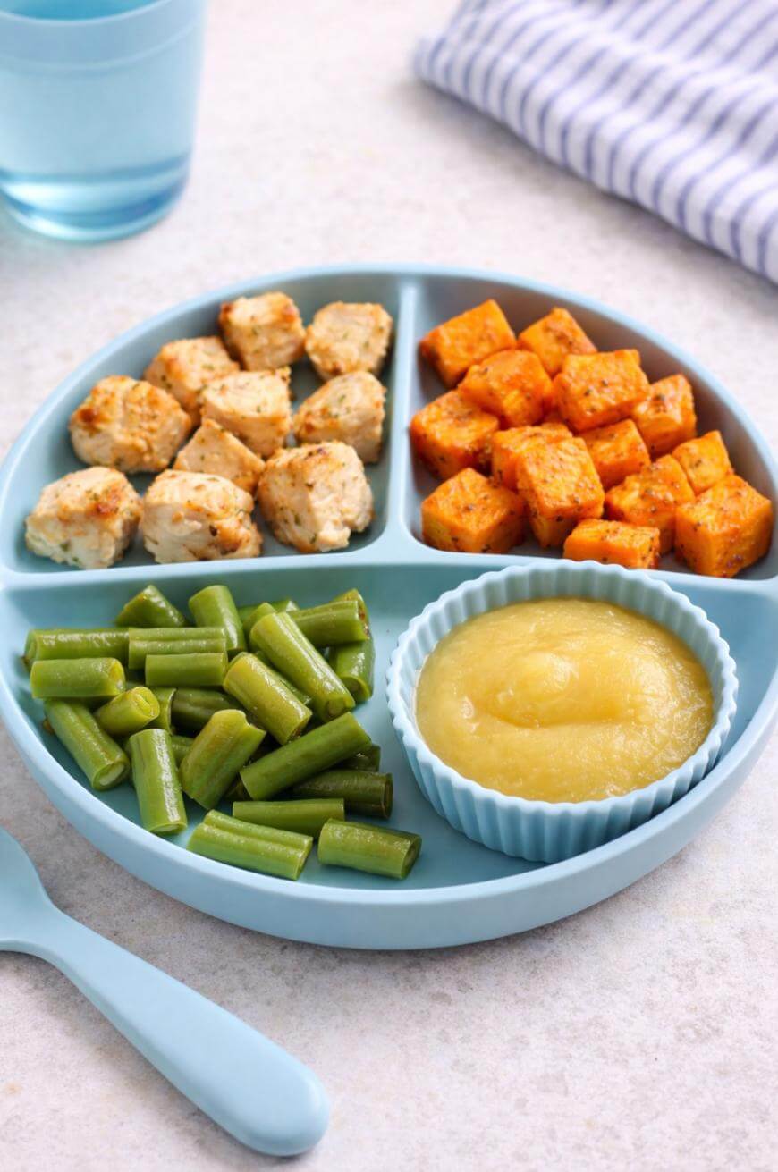 A blue divided plate with sections containing diced chicken, roasted sweet potato cubes, cut green beans, and a silicone cup filled with applesauce. A blue spoon and a glass of water are nearby.