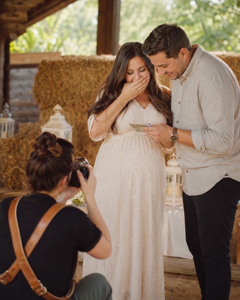 A pregnant woman in a white dress covers her mouth in surprise while a man beside her smiles at a note in his hand. A photographer kneels in front of them, capturing the joyful moment indoors near hay bales.