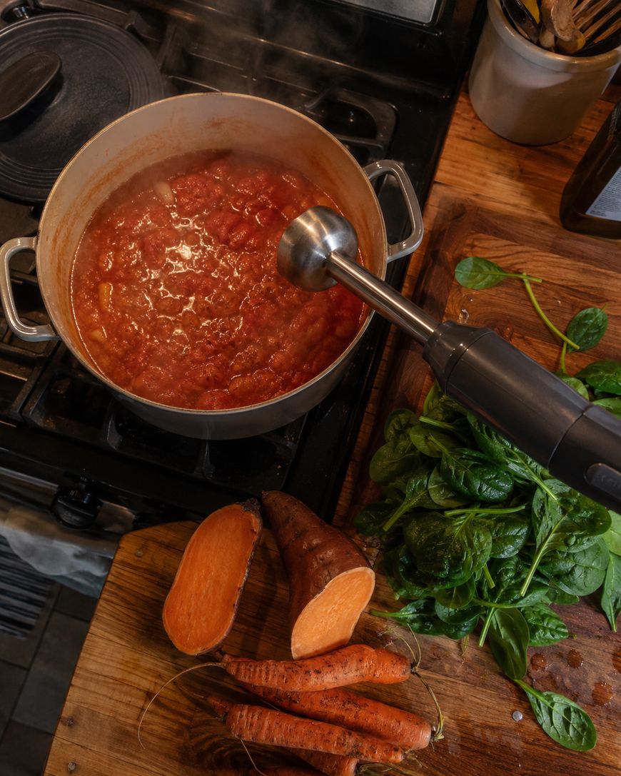 A pot of tomato sauce cooking on a stove is being blended with an immersion blender. Fresh spinach, sweet potatoes, and carrots are on a wooden cutting board nearby. Steam rises from the pot.