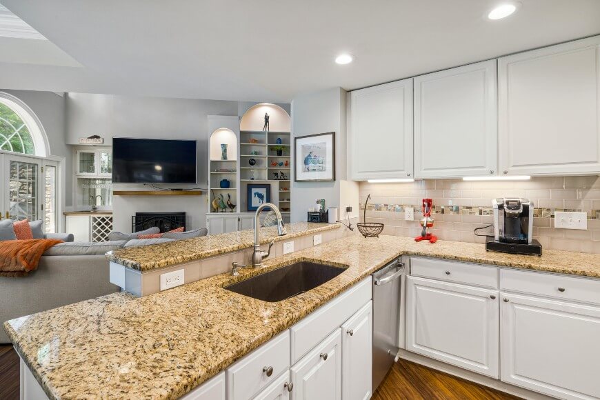 A modern kitchen with granite countertops, white cabinets, and a stainless steel sink and dishwasher. The open-concept layout connects to a living area with a fireplace and TV.