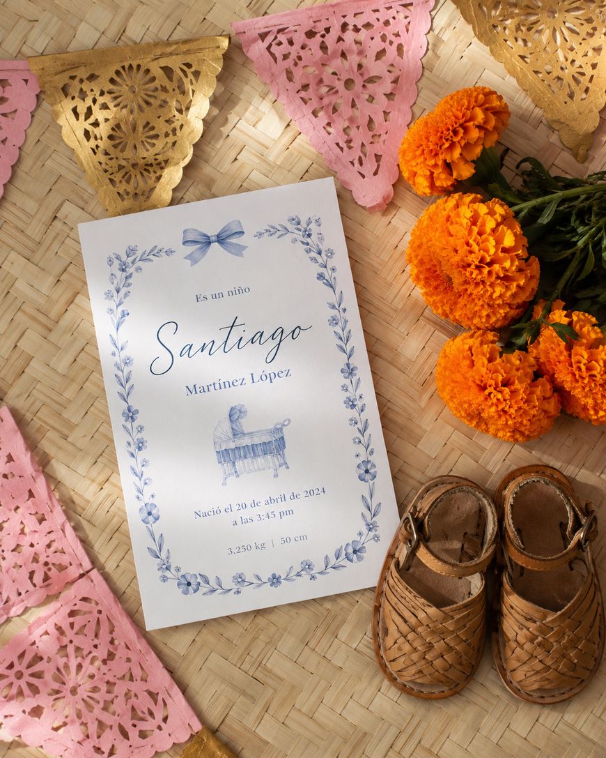 A birth announcement card for Santiago lies on a woven mat with orange marigolds, woven baby sandals, and pink and gold papel picado decorations around it. The card has blue floral designs and baby details in Spanish.