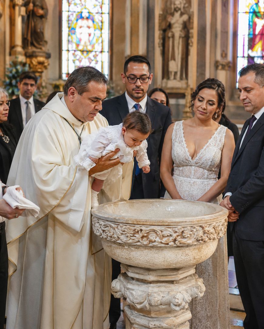 A priest holds a baby over a baptismal font during a baptism ceremony in a church, with the baby’s parents and others standing around, watching attentively. Stained glass windows and ornate decorations are visible in the background.