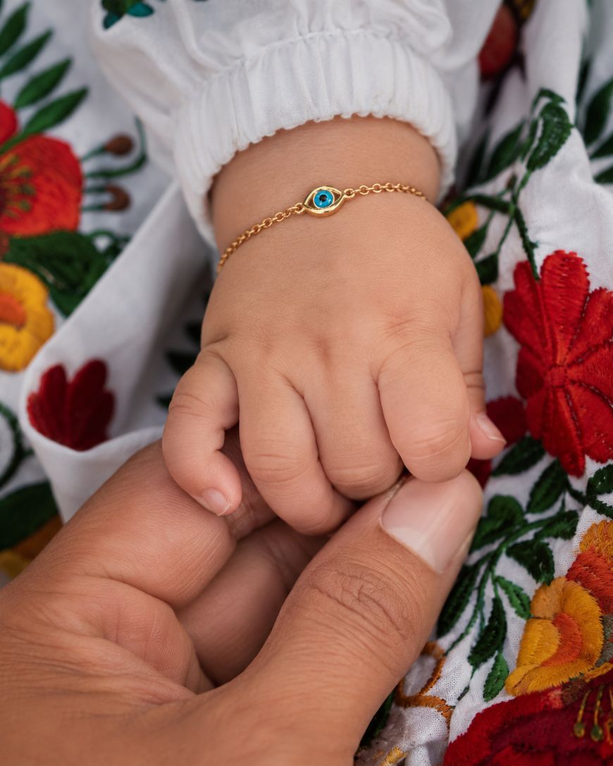 A baby’s hand wearing a gold bracelet with a blue charm holds an adult’s finger. Both hands rest on white clothing decorated with colorful embroidered flowers.