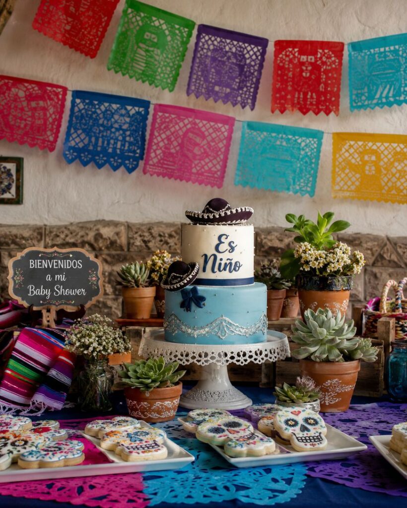 A baby shower table decorated with colorful papel picado, succulents, sugar skull cookies, and a blue cake that says Es Niño. A sign reads Bienvenidos a mi Baby Shower in Spanish.